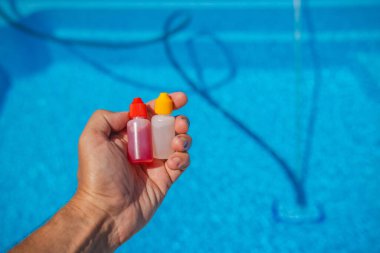 Güneş ışığında temiz mavi bir yüzme havuzunun üzerinde havuz suyu testi için kullanılan kırmızı ve sarı şapkalı iki damlatıcıyı tutan elin yakın çekimi. Yüksek kalite fotoğraf