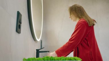 Kırmızı bornozlu genç bir kadın modern bir banyoda elleri akan suyun altında yuvarlak ışıklı bir aynayla yıkıyor. Yüksek kalite 4k görüntü