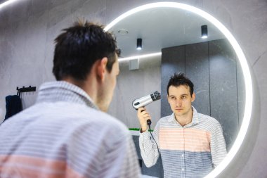 Islak saçlı genç bir adam modern bir banyoda aynaya bakarken saç kurutma makinesini tutuyor ve sabah bakım rutinine odaklanıyor. Yüksek kalite fotoğraf