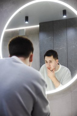 Genç adam endişeli bir bakışla aynada yüzünü inceliyor, modern bir banyoda sabah rutini sırasında nazikçe çenesine dokunuyor. Yüksek kalite fotoğraf