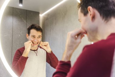 Genç bir adam, dişlerini aynanın önünde diş ipiyle temizlerken günlük kendine bakımı sırasında hafifçe gülümsüyor. Yüksek kalite fotoğraf