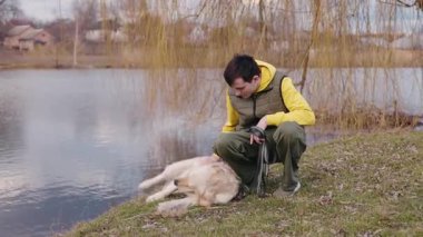 Genç adam, söğüt dallarının altındaki sakin bir gölün yakınındaki çimlerde uzanan Golden Retriever 'ını şefkatle okşar. Yüksek kalite 4k görüntü