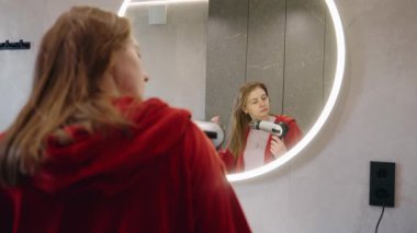 Kırmızı bornozlu genç kız saçlarını yuvarlak aynanın yanında saç kurutma makinesiyle kurutuyor. Yüksek kalite 4k görüntü