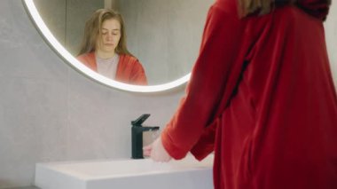Kırmızı bornozlu genç kız banyo aynasında kendine bakarken musluğun altında ellerini yıkıyor. Yüksek kalite 4k görüntü