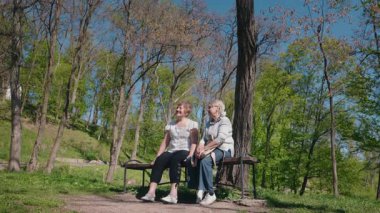 İki yaşlı kadın güneşli bir orman parkında bir bankta oturuyor, taze bahar havasında gülümsüyor ve sohbet ediyorlar. Yüksek kalite 4k görüntü