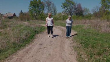 Güneşli bir günde kırsal kesimde, iki yaşlı kadın toprak yolda birlikte yürürken, içlerinden biri bacak ameliyatı nedeniyle topallayarak yavaşça yürüyor. Yüksek kalite 4k görüntü