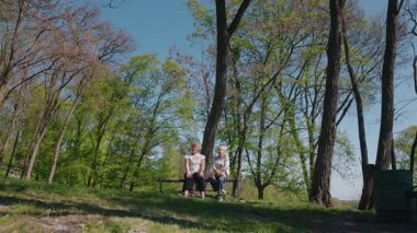 İki son sınıf öğrencisi kadın, ağaçlar ve güneş ışığıyla çevrili bir bahar ormanındaki bankta neşeli bir sohbetin tadını çıkarıyorlar. Yüksek kalite 4k görüntü