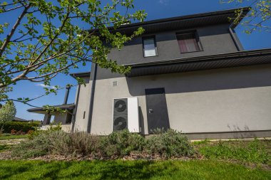 Çağdaş bir evin dış duvarına, taze çimenler, çalılar ve sürdürülebilir bir bahçe ortamında çiçek açan ağaçlarla çevrili, enerji tasarruflu bir ısı pompası.