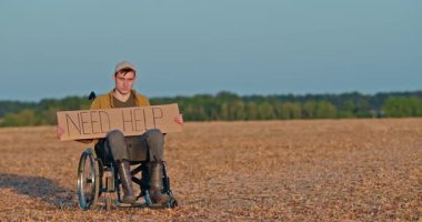 Tekerlekli sandalyedeki genç adam açık gökyüzünün altındaki kuru bir arazide tek başına otururken yardıma ihtiyacın var yazan bir karton tabela tutuyor. Yüksek kalite 4k görüntü