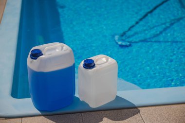 Mavi kapaklı iki plastik kimyasal kap, parlak güneş ışığında su arıtma ve bakım için kullanılan bir yüzme havuzunun kenarına yerleştirilmiş. Yüksek kalite fotoğraf