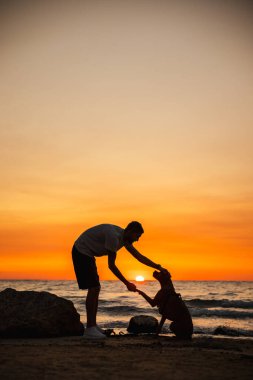 Adam, canlı bir günbatımında patisini kumlu bir sahilde tutarken Alman boksör köpeğini okşuyor. Arka planda okyanus dalgaları olan siluet bir manzara. Yüksek kalite fotoğraf
