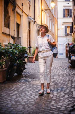 Orta yaşlı, açık renk elbiseli ve hasır şapkalı bir kadın Roma 'da dar bir kaldırım taşı caddesinde yürüyor ve gece lambalarıyla aydınlatılmış tarihi mimariye bakıyor. Yüksek kalite fotoğraf
