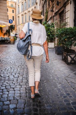 Orta yaşlı, hasır şapkalı ve yaz kıyafetli bir kadın sırt çantasıyla Roma 'da bir kaldırım taşı caddesinde yürüyor. Yüksek kalite fotoğraf