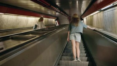 İki kadın metro istasyonunda yürüyen merdivene biniyor, biri şapka ve günlük kıyafet içinde, diğeri ise sırt çantalı beyaz bir elbise içinde. Yüksek kalite 4k görüntü
