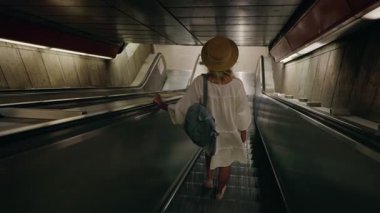 Beyaz elbiseli, sırt çantalı genç bir kadın metro istasyonunda yürüyen merdivenden iniyor. Yüksek kalite 4k görüntü