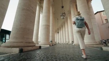 Sırt çantalı orta yaşlı bir kadın Vatikan 'daki St. Peters Meydanı' nın büyük taş sütununun altında yürüyor..