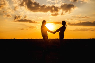 Gün batımında bir tarlada el ele tutuşan iki kadın, altın gökyüzüne karşı siluette yakalandılar. Yüksek kalite fotoğraf