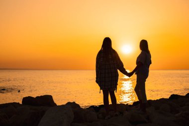 Gün doğumunda kayalık sahilde el ele tutuşan anne ve kızın silueti aile bağını, duygusal yakınlığı, refahı, güveni ve huzur dolu bir deniz kenarı yaşam tarzını simgeliyor. Yüksek kalite fotoğraf