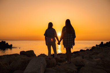 Gün doğumunda deniz kenarındaki kayalarda el ele tutuşan iki kadının silueti. Yüksek kalite fotoğraf