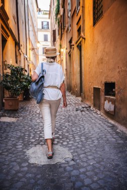 Orta yaşlı, açık renk elbiseli ve hasır şapkalı bir kadın Roma 'da eski binalarla çevrili dar bir kaldırım taşı sokakta sırt çantasıyla yürüyor. Yüksek kalite fotoğraf