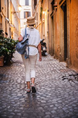 Eski duvarlarında sıcak akşam ışığı olan dar bir Roma geçidinde yürürken orta yaşlı bir kadın sırt çantası ve hasır şapka taşıyor. Yüksek kalite fotoğraf