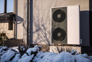 Modern hava kaynağı ısı pompası kışın bir yerleşim yerinin dışına kurulur. Karla kaplı bitkiler ve güneş ışığı sürdürülebilir ev ısıtma ve enerji verimliliği kavramını yansıtıyor. Yüksek kalite