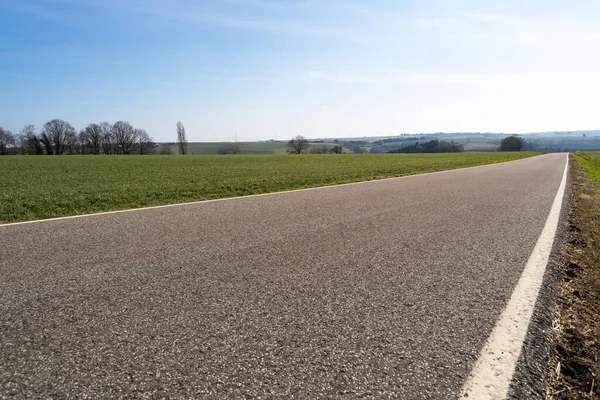 Low angle view of a paved rural road between grassfields in the landscape with blue sunny sky in spring