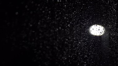 Unfocused raindrops run down a window at night while it's raining and a street lamp is glow in the background