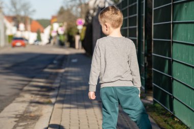 Five year old boy wearing a gray sweater and green sweatpants is standing on the sidewalk next to a fence