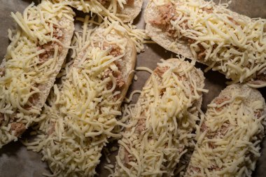 Top view of many unbaked baguette buns with tuna and cheese on a baking sheet