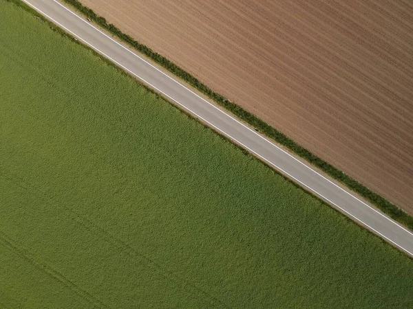 从一条柏油路的上方眺望 这条柏油路介于一个绿色生长的农田和一个有泥土的褐色耕地之间 — 图库照片