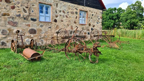 Old antique farm tools are displayed on the green grass outside a rustic stone barn.