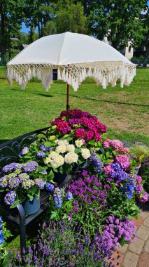 Bahçede beyaz püsküllü şemsiye ve metal bankın etrafında bir sürü güzel çiçek..