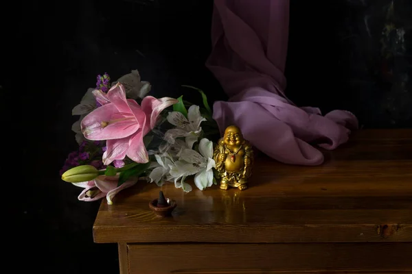 Still life with a figure of a laughing buddha, a bouquet of flowers and a smoking incense close-up on a bedside table
