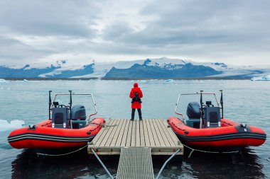 A tourist in a red jacket stands on a pier and looks at the snow-capped mountains. Iceland. Two red motor boats are moored near the pier.