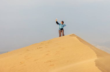 Dişi bir turist kum tepesinin tepesinde durur ve selfie çeker. Huacachina Çölü. Ica bölgesi. Peru