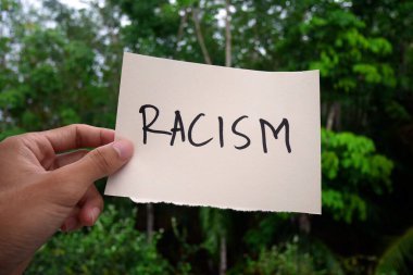 Hand holding white Paper with racism text in solative on nature background. Racism concept. Discrimination, racial issues clipart