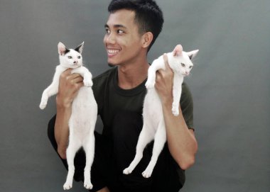 A young Asian man holds two white kittens, one in each hand. his eyes widened with curiosity. The man's face lit up with a warm, gentle smile. clipart