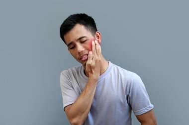 Genç Endonezyalı bir adam çenesini acılı bir ifadeyle tutuyor, bu da ciddi diş ağrısı olduğunu gösteriyor.