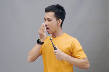 A young Asi man in a yellow t-shirt stands on a gray background, holding an electric toothbrush. He covered his mouth with his hand and looked disgusted by the problem of bad breath clipart