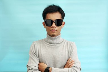 Balıkçı yaka kazak giyen, güneş gözlüğü takan ve kollarını kavuşturan yakışıklı Asyalı genç adamın portresi.