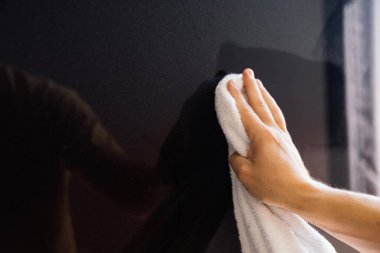 Beyaz bezle plazma paneline toz silen bir adam. Bir el beyaz bir bez ile parlak siyah bir yüzeyi silerken görülüyor, iyi aydınlatılmış bir kapalı alanda temizliği sürdürme çabasını vurguluyor..