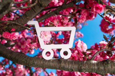 Beyaz alışveriş arabası ikonu çiçek açan kiraz çiçeklerinin arasına yerleştirilmiş parlak pembe çiçekli ve mavi gökyüzü.