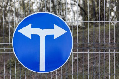 Mavi dairesel yol işareti, arka planda ağaçlık bir alan olan tel örgüye monte edilmiş, zorunlu sola ya da sağa dönüş olduğunu gösteriyor..