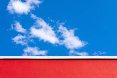 Kırmızı bir duvarın minimalist görüntüsü, beyaz süslemeli, parlak mavi gökyüzüne karşı dağınık bulutlu..
