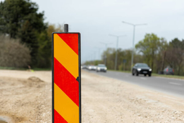Close-up of a red and yellow diagonal striped roadwork sign on a roadside under cloudy sky with traffic in the background.