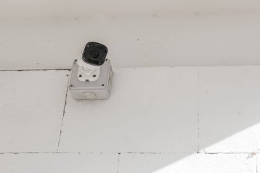 Güvenlik kamerası beyaz fayanslı bir duvara monte edilmiş. Hafif tozlu ve parlak bir ortamda tepeden izlenmek üzere yerleştirilmiş..