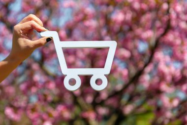 Güneşli bir günde arka planda pembe kiraz çiçeklerinin açtığı beyaz bir alışveriş arabası sembolü..