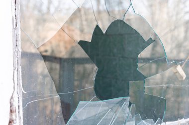 Çentikli cam kırıkları ve büyük bir deliği olan kırık bir pencerenin yakın çekimi, terk edilmiş bir iç mekan ve dış mekanın yansımalarını ortaya çıkarıyor..