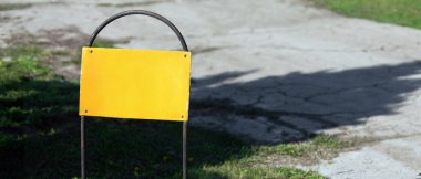 Güneşli bir parkın çatlamış kaldırımının yanındaki çimlerin üzerinde duran metal çerçevede yıpranmış boş sarı bir tabelanın yakın çekimi.